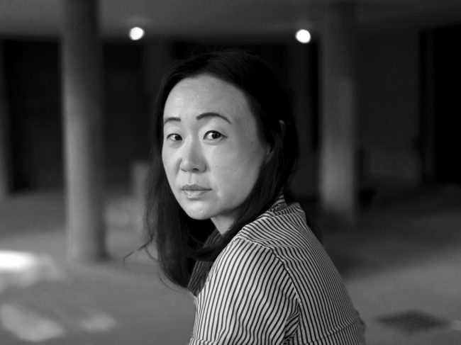 A black-and-white profile photo of Sandra Lim in an empty gallery space and looking toward the camera over her left shoulder.