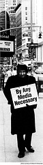 Man on sidewalk holding sign reading "By Any Media Necessary"