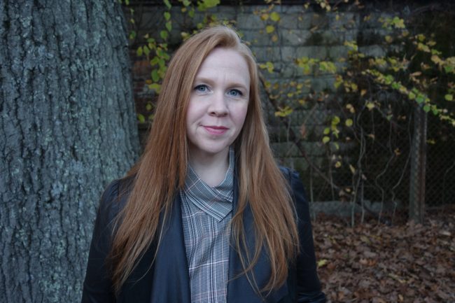 Alison Adair with long red hair and standing in front of a tree and garden wall.