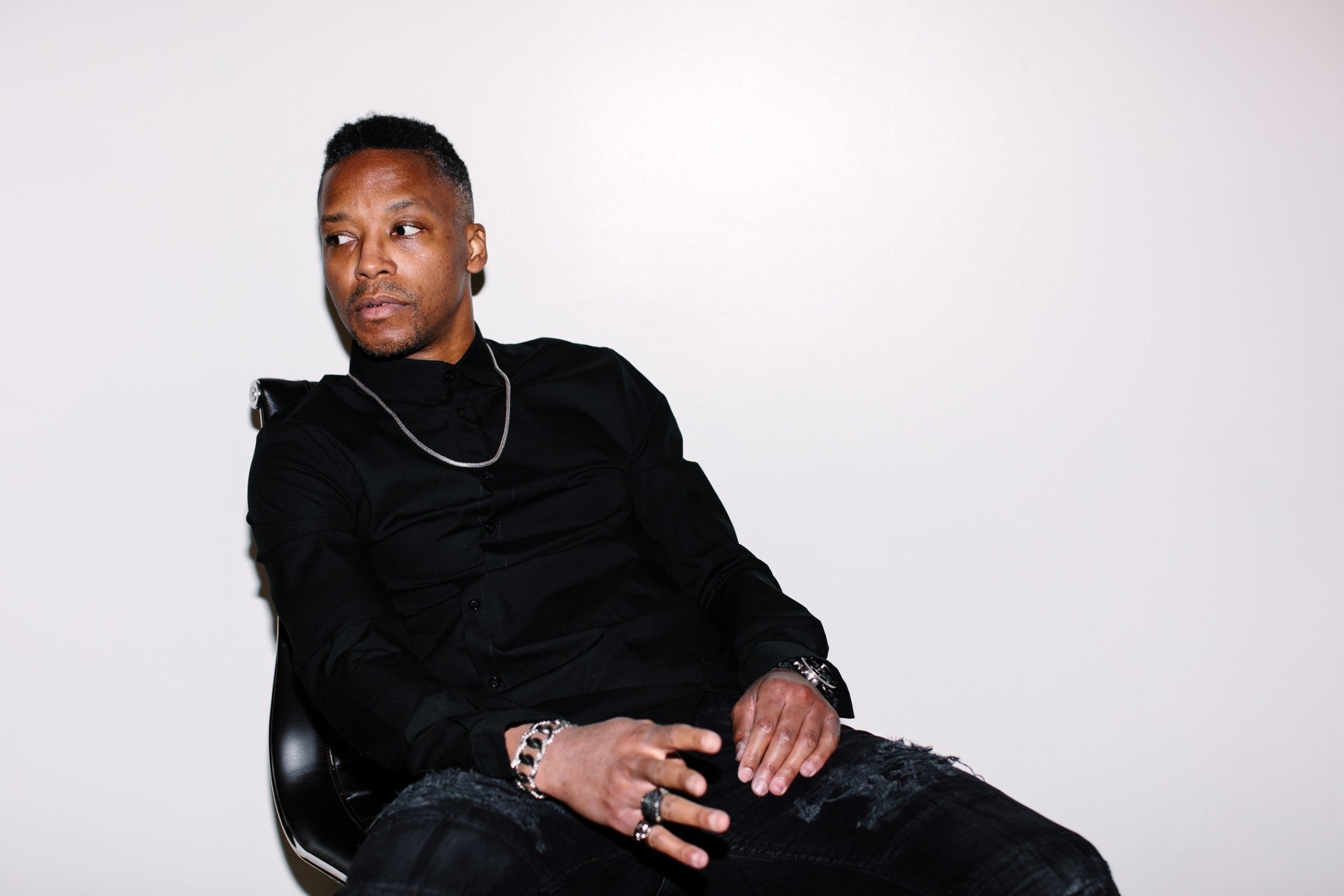 Lupe Fiasco sitting in a chair in front of a bright wall. He is wearing all-black and a silver necklace.