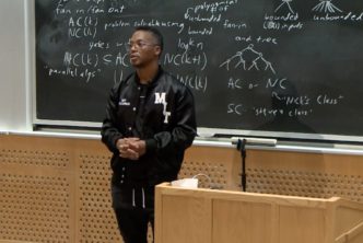 Lupe Fiasco presenting in front of a lecturn and wearing a black jacket with the letters MIT on it in white