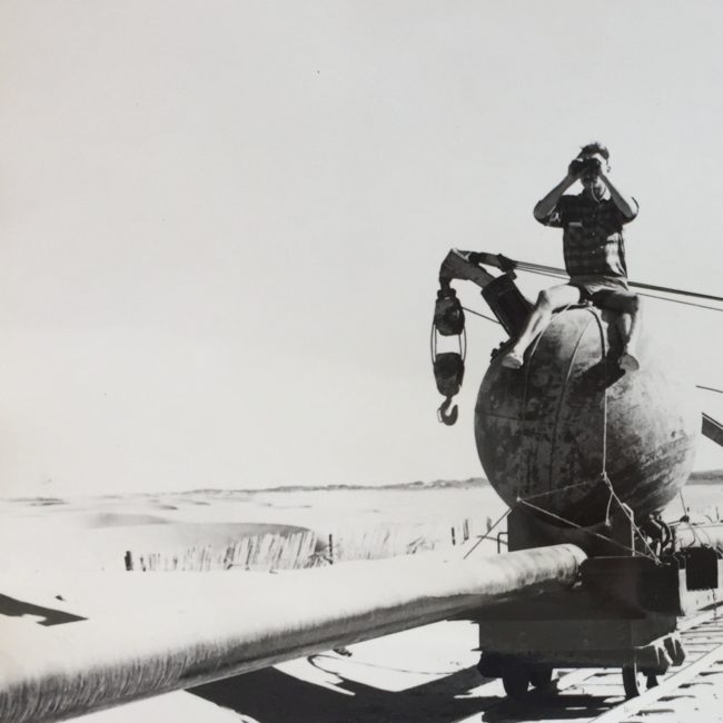 Black and white photo of a man with binoculars sitting on a wrecking ball. The wrecking ball is on a small railcar on tracks and holding up a pipline.