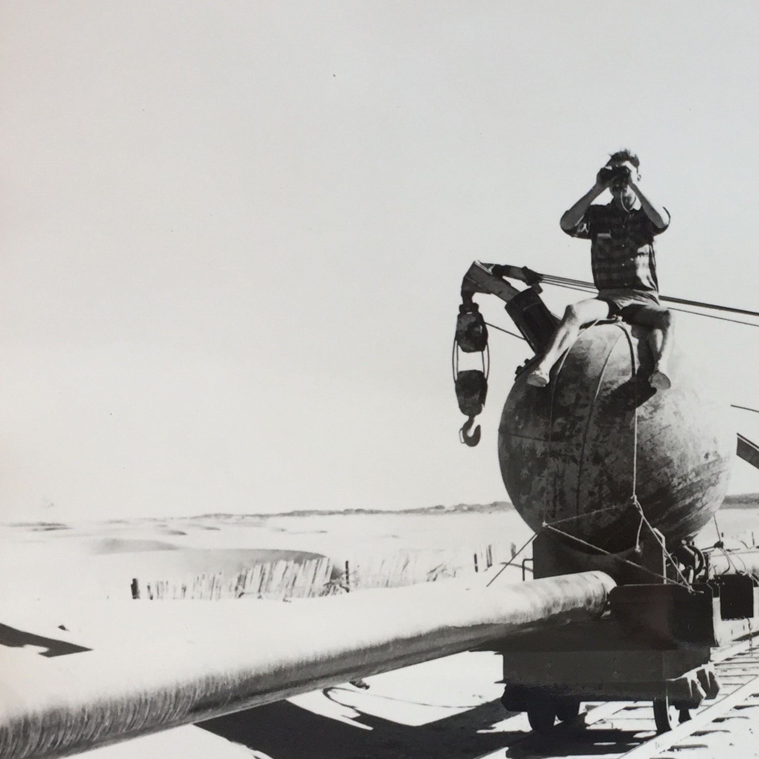 Black and white photo of a man with binoculars sitting on a wrecking ball. The wrecking ball is on a small railcar on tracks and holding up a pipline.